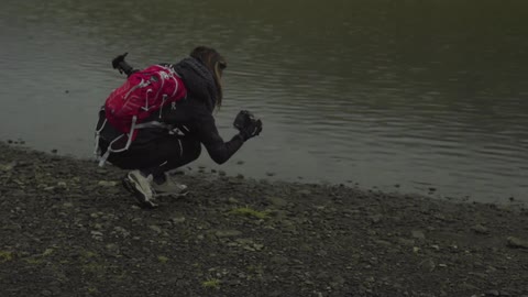 A woman wearing a pink backpack squatting in front of a body of water while looking at a camera