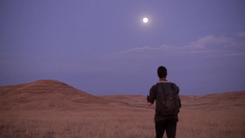A man wearing dark clothing walks across a sandy desert toward distant mountains