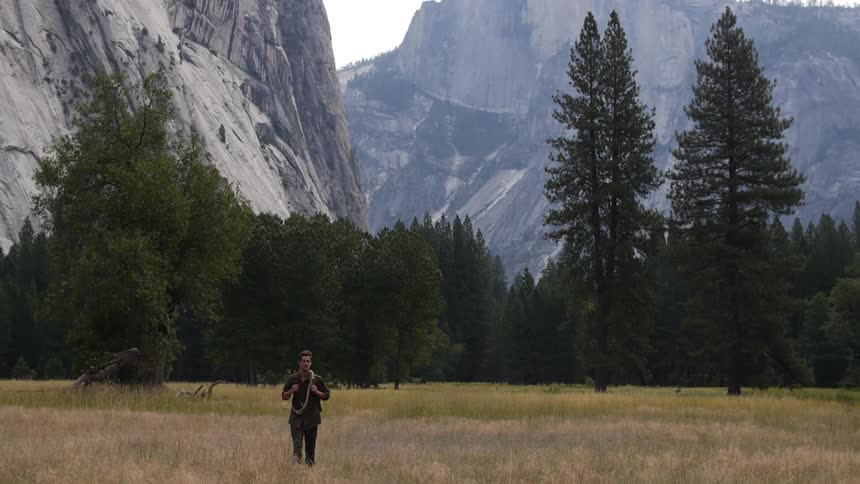A man walking through grass in front of large snow covered mountains with green trees
