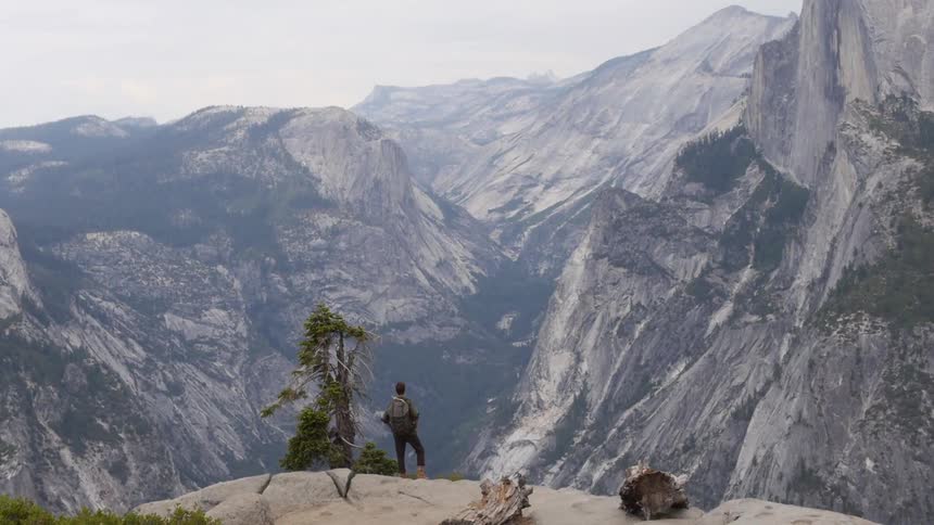 A person stands on a cliff looking out at large mountains on a cloudy day