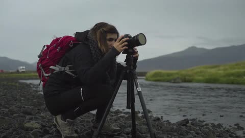 A squatting woman takes photos of a small river using a camera mounted on a tripod