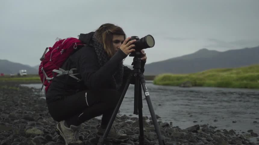 A squatting woman takes photos of a small river using a camera mounted on a tripod