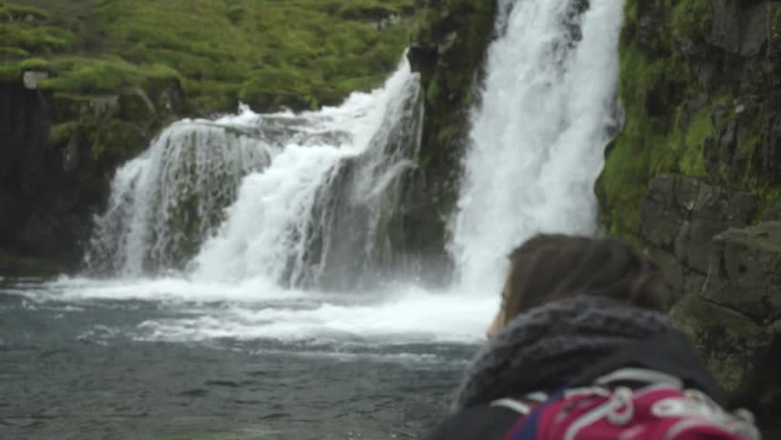 A woman with a pink backpack walks towards a waterfall