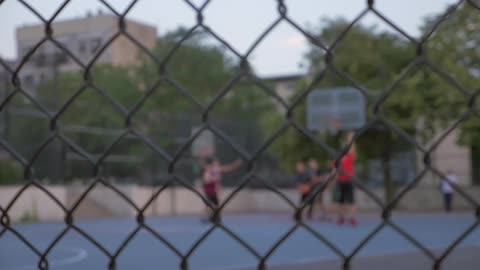 A group of boys play basketball on a fenced in court in the daytime