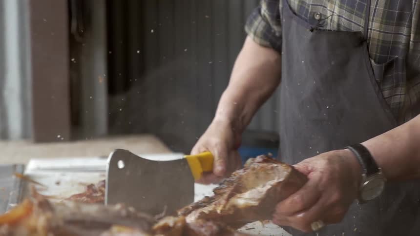 A man chops smoking barbecued ribs on a countertop near a grill