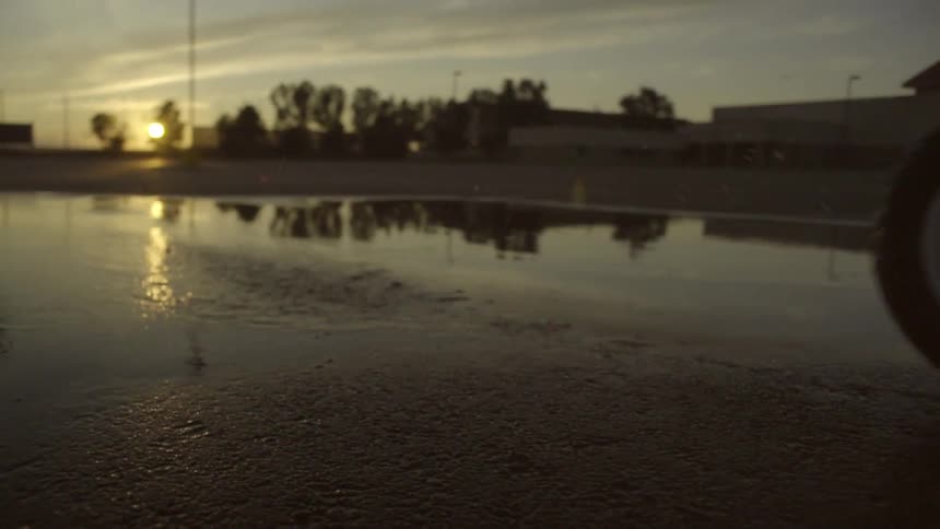 Two children wearinghelmets are riding their bicycles though a puddle while a dog watches and the sun sets in the background