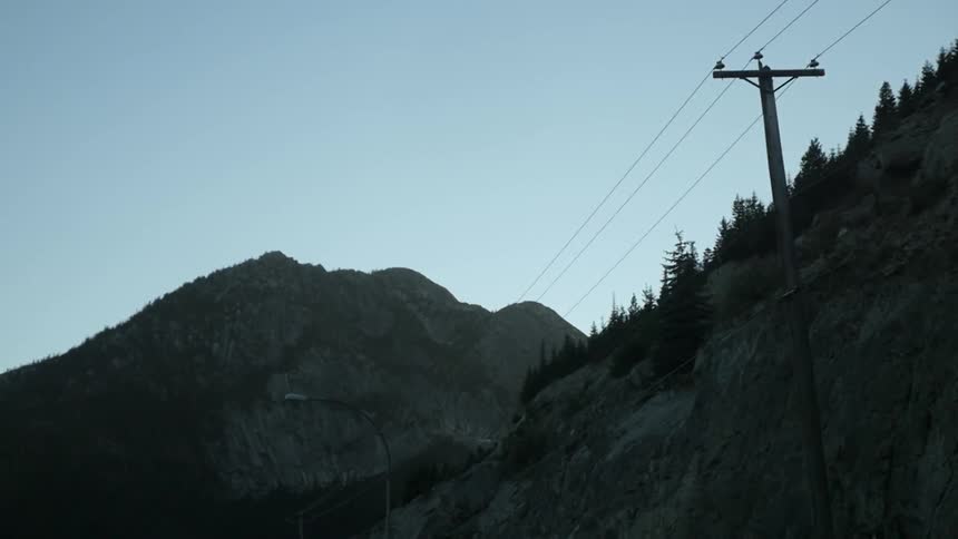 Rocky mountains lined with trees next to a road with street lines and power lines