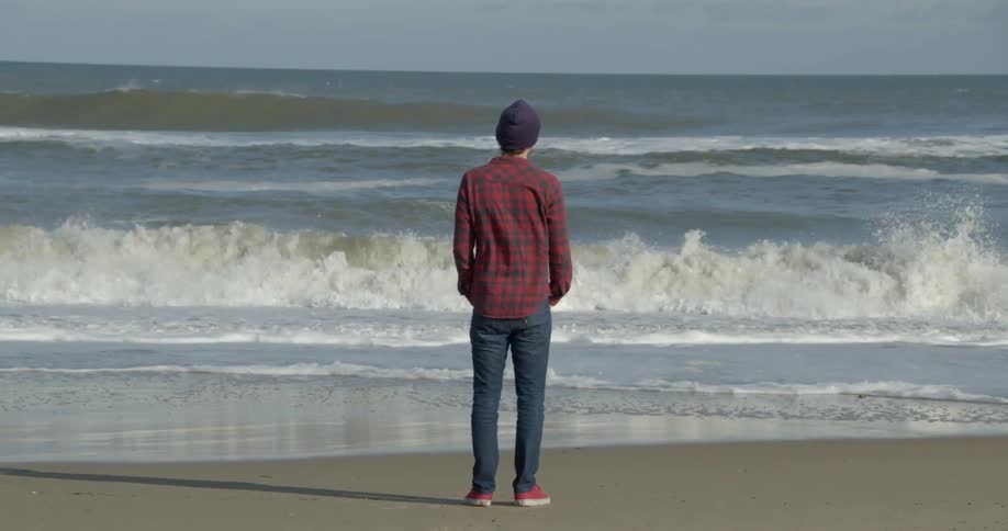 A man standing at shore and watching the waves crash with a grey sky