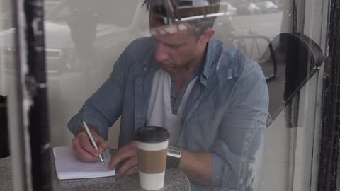 A man sits and writes at a table with a cup of coffee
