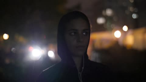 A woman stands in the city at night listening to music on her headphones and nodding her head