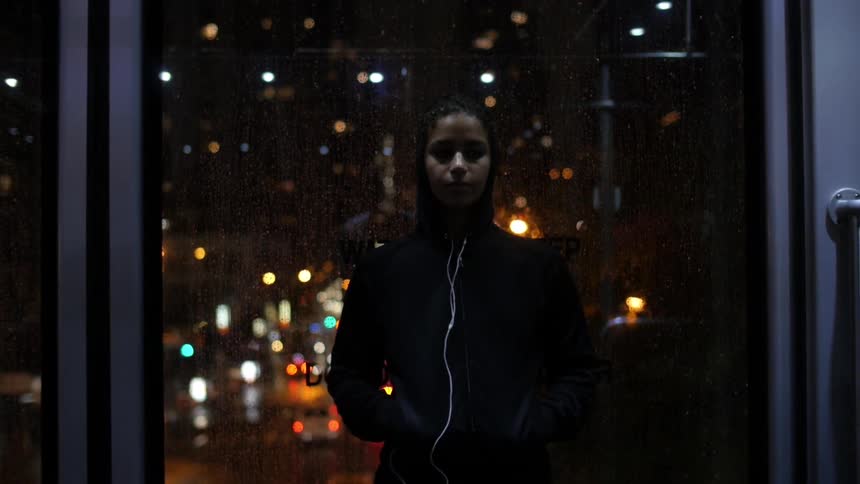 A woman stands by a window in the city on a rainy night