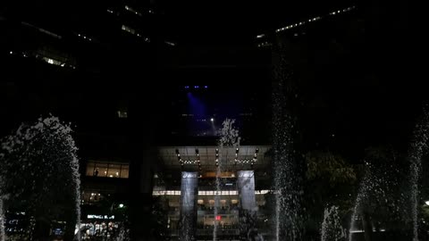 A water with multiple spouts is displayed in front of a city building