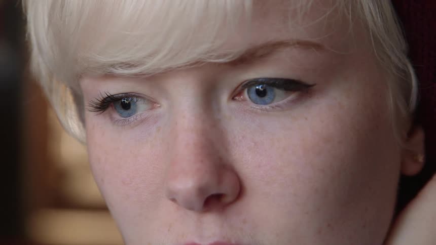 A close up of a young blonde woman with blue eyes watching something before looking down and away