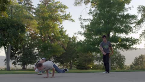 A group of young men break dancing in a park with trees behind them on a sunny day