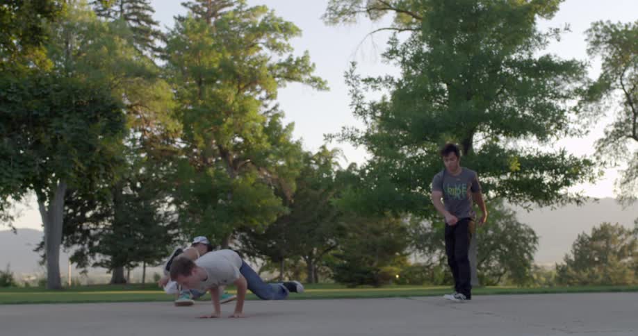 A group of young men break dancing in a park with trees behind them on a sunny day