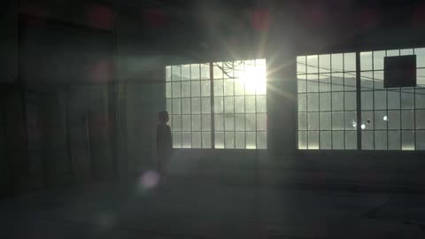 A person stands in an empty warehouse and stares out the large windows