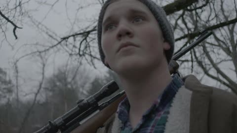 A young man stands holding a gun over his shoulder in the woods