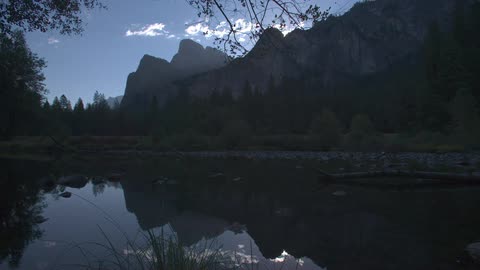 A still body of water surrounded by trees at the bottom of mountains with a blue sky and a few little white clouds