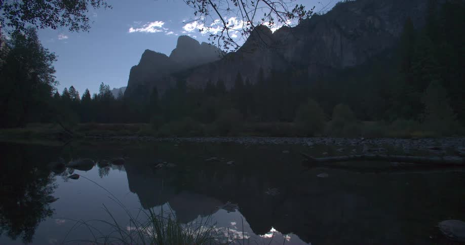 A still body of water surrounded by trees at the bottom of mountains with a blue sky and a few little white clouds