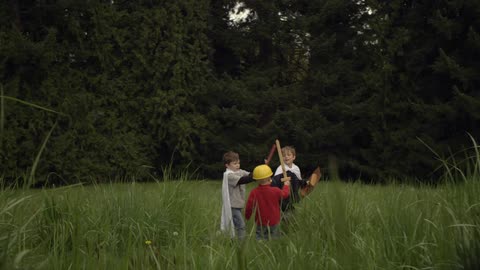 Children dressed in makeshift capes playing with wooden swords in a field that is surrounded by trees