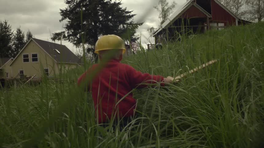 A little boy in a yellow hat holding a wooden sword stumbles through the tall grass