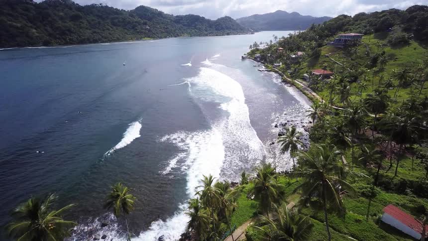 Waves crashing onto a lush, tropical beach surrounded by hills and a few homes on a sunny day