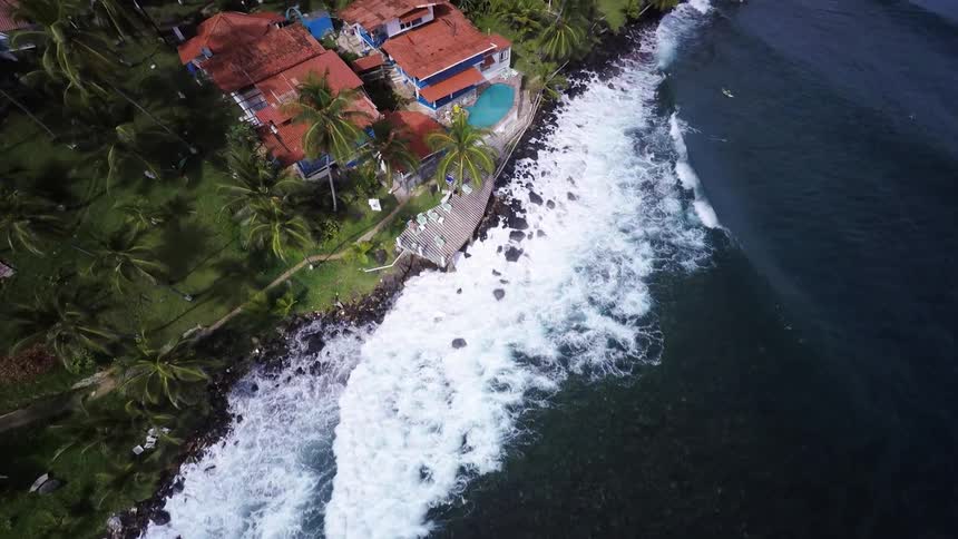 Waves crash on a rocky beach right next to some buildings