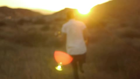 A man running through tall grasses towards hills that the sun is setting behind